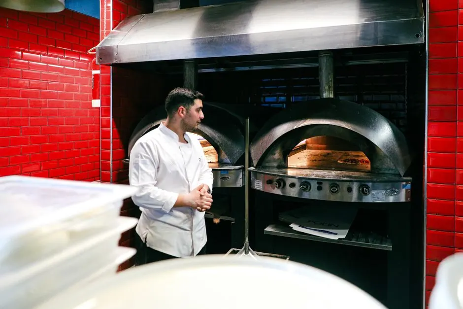 Chef in white uniform stands by pizza ovens in a vibrant restaurant kitchen, ready to bake.